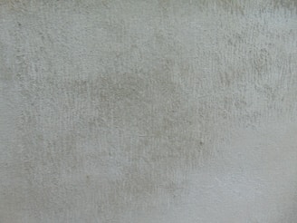 A textured surface with a rough, uneven look, featuring subtle variations in color and depth. The texture resembles old concrete or plaster with visible marks and irregularities.