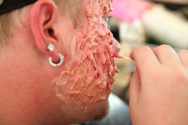A close-up of a person's cheek with makeup or prosthetics applied to create a textured, fleshy appearance with shades of red and pink. The image includes part of the person's ear with a small gauge earring, and a hand is partially visible applying the makeup with a small brush.