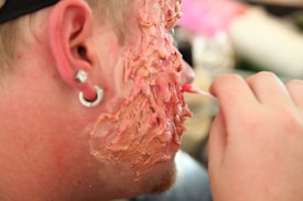A close-up of a person's cheek with makeup or prosthetics applied to create a textured, fleshy appearance with shades of red and pink. The image includes part of the person's ear with a small gauge earring, and a hand is partially visible applying the makeup with a small brush.