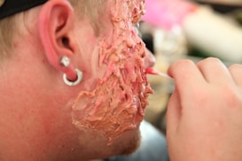 A close-up of a person's cheek with makeup or prosthetics applied to create a textured, fleshy appearance with shades of red and pink. The image includes part of the person's ear with a small gauge earring, and a hand is partially visible applying the makeup with a small brush.