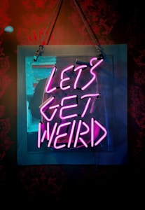 A neon sign with bold, brightly lit pink letters that reads 'LET'S GET WEIRD', hanging on a wall. The sign is rectangular, transparent, and the light emanates a vibrant glow against the dark, subtly patterned background.