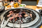 A sizzling barbecue grill with pieces of marbled meat cooking over glowing coals. The grill is set on a dining table with blurred background elements including drinks and side dishes.