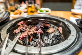 A sizzling barbecue grill with pieces of marbled meat cooking over glowing coals. The grill is set on a dining table with blurred background elements including drinks and side dishes.
