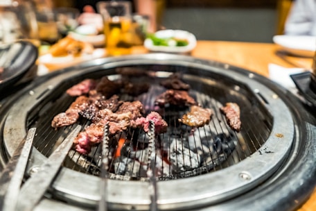 A sizzling barbecue grill with pieces of marbled meat cooking over glowing coals. The grill is set on a dining table with blurred background elements including drinks and side dishes.