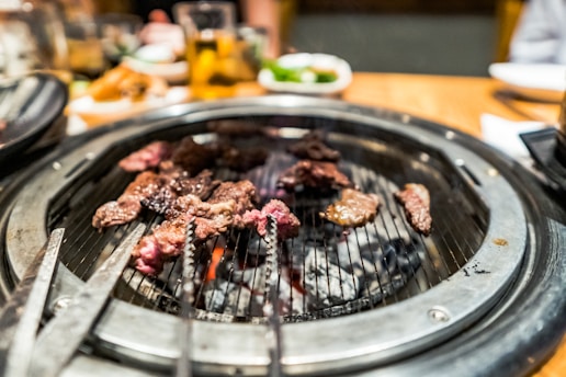 A sizzling barbecue grill with pieces of marbled meat cooking over glowing coals. The grill is set on a dining table with blurred background elements including drinks and side dishes.