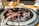 A sizzling barbecue grill with pieces of marbled meat cooking over glowing coals. The grill is set on a dining table with blurred background elements including drinks and side dishes.