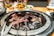 A sizzling barbecue grill with pieces of marbled meat cooking over glowing coals. The grill is set on a dining table with blurred background elements including drinks and side dishes.
