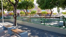 A playground surrounded by a fence, featuring various play equipment such as swings and climbing structures. There are benches along the edge, shaded by large trees. In the background, a colorful building with decorative features and cars parked along the street can be seen.