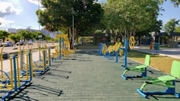 An outdoor fitness area featuring a variety of exercise equipment, including stationary bikes and other workout stations. The equipment is primarily blue and yellow, arranged neatly on a paved surface. Surrounding the area are trees providing shade, with a few parked cars and buildings visible in the background.