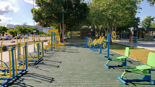 An outdoor fitness area featuring a variety of exercise equipment, including stationary bikes and other workout stations. The equipment is primarily blue and yellow, arranged neatly on a paved surface. Surrounding the area are trees providing shade, with a few parked cars and buildings visible in the background.