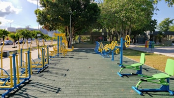 An outdoor fitness area featuring a variety of exercise equipment, including stationary bikes and other workout stations. The equipment is primarily blue and yellow, arranged neatly on a paved surface. Surrounding the area are trees providing shade, with a few parked cars and buildings visible in the background.