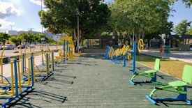 An outdoor fitness area featuring a variety of exercise equipment, including stationary bikes and other workout stations. The equipment is primarily blue and yellow, arranged neatly on a paved surface. Surrounding the area are trees providing shade, with a few parked cars and buildings visible in the background.