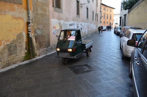 A narrow cobblestone street lined with parked cars on the right and rustic buildings on the left. A small, green three-wheeled vehicle is driving down the street, with a for-sale sign in its window. Several pedestrians are visible in the background, walking further down the street. The walls of the buildings are adorned with faded paint and graffiti.