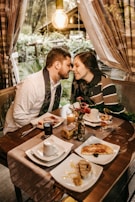 A couple lovingly leans toward each other over a table set for a meal. The table is decorated with various breakfast items, including coffee, juice, toast, and pastries. A small vase with red flowers and a lit candle add to the cozy atmosphere. The setting is indoors with a large window providing a view of a lush, green garden. Warm lighting from an exposed bulb creates an intimate and inviting ambiance.