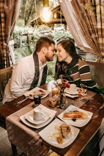 A couple lovingly leans toward each other over a table set for a meal. The table is decorated with various breakfast items, including coffee, juice, toast, and pastries. A small vase with red flowers and a lit candle add to the cozy atmosphere. The setting is indoors with a large window providing a view of a lush, green garden. Warm lighting from an exposed bulb creates an intimate and inviting ambiance.
