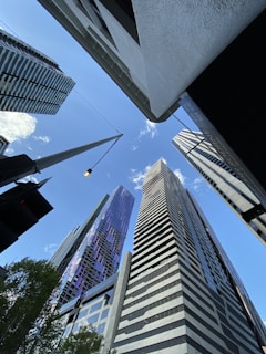 A group of modern skyscrapers viewed from below against a clear blue sky. The buildings are tall and architecturally diverse, with reflective glass surfaces and varying geometric patterns. A streetlight is visible in the foreground, and there are hints of greenery from trees at the bottom edge.