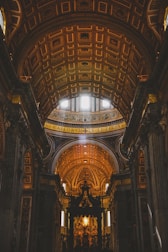 An ornate, grand cathedral interior is depicted, featuring intricate architectural details and a high, arched ceiling adorned with elaborate patterns. Golden tones dominate, with light streaming through small windows, illuminating the rich decorations.