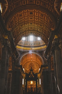 An ornate, grand cathedral interior is depicted, featuring intricate architectural details and a high, arched ceiling adorned with elaborate patterns. Golden tones dominate, with light streaming through small windows, illuminating the rich decorations.