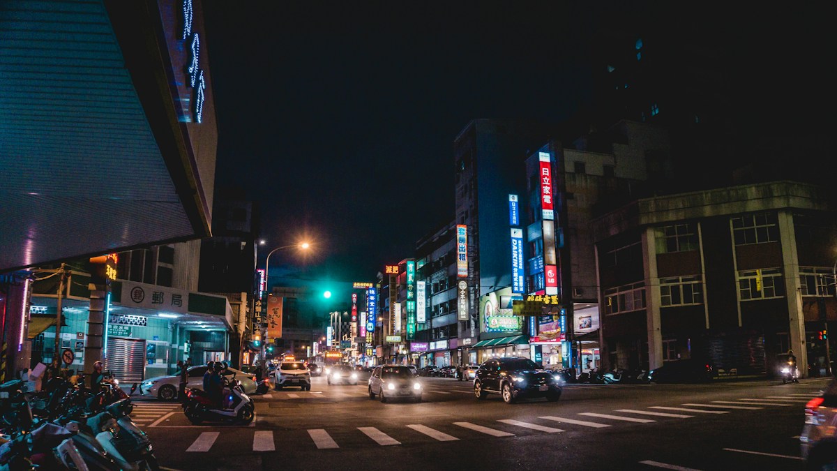 A city intersection in Taiwan at night, with scooters at the corner and cars crossing the zebra crossing