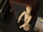 A wooden artist mannequin is positioned near a speaker and a CD case on a dark surface. The mannequin, made of smooth, light-colored wood, stands upright with slightly bent limbs. The speaker has a dark fabric front with a wooden frame, while the CD case displays text and graphics related to music.