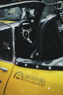 A close-up view of a yellow sports car interior, focusing on the steering wheel and partial dashboard. The steering wheel features a three-spoke design, and there is a metallic finish with buttons or controls around the dashboard. The car's frame and seat are also visible, with a leather or leather-like material.