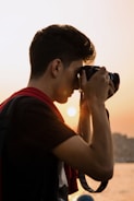 A person is holding a camera up to their face, capturing a picture at sunset. The sun creates a warm glow in the background, and the individual appears focused on their photography.