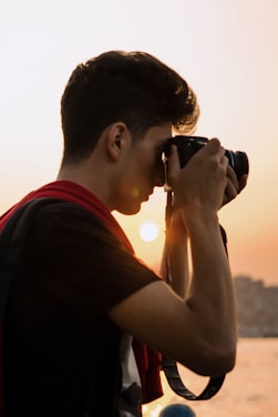 A person is holding a camera up to their face, capturing a picture at sunset. The sun creates a warm glow in the background, and the individual appears focused on their photography.