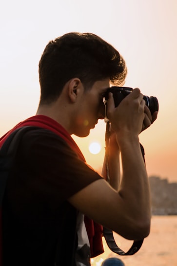 A person is holding a camera up to their face, capturing a picture at sunset. The sun creates a warm glow in the background, and the individual appears focused on their photography.