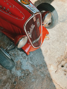A vintage red car with chrome details and a classic front grille is parked on a rough concrete surface. The car features distinctive headlamps and a visible emblem with the word 'Balilla'. The vehicle is captured under natural light, casting shadows on the ground.