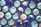 Rows of glass marbles in various shades of blue and turquoise are arranged against a textured gray surface. Some marbles show reflections of light that create hints of other colors, adding to their glossy appearance.
