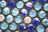 Rows of glass marbles in various shades of blue and turquoise are arranged against a textured gray surface. Some marbles show reflections of light that create hints of other colors, adding to their glossy appearance.