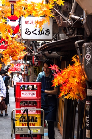 A narrow alley adorned with vibrant autumn leaves is bustling with activity. A person in a blue apron maneuvers a cart filled with red crates labeled 'Sapporo' and 'Kirin,' suggesting beer bottles inside. Various signs with Japanese characters hang above, creating a lively and colorful atmosphere.