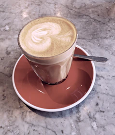 A hand holding one of our elegant coffee spoons, poised over a frothy latte in a cozy café setting.