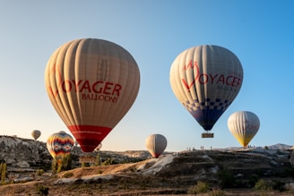 Several hot air balloons are floating in the sky above a rocky landscape. The balloons are large, with the words 'Voyager Balloons' printed on them. The landscape below features rugged terrain with sparse vegetation and small hills.