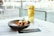 A glass of Heineken beer sits next to a black bowl filled with chicken wings and a dipping sauce on a table. The setting appears to be a cafe or restaurant with large glass windows overlooking an outdoor area.