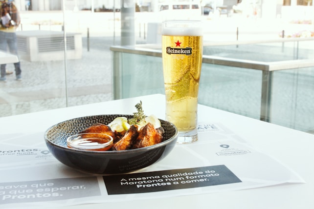 A glass of Heineken beer sits next to a black bowl filled with chicken wings and a dipping sauce on a table. The setting appears to be a cafe or restaurant with large glass windows overlooking an outdoor area.