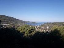 A scenic coastal landscape with a view of hills covered in dense greenery, a small village with red-roofed houses near the coast, and a calm sea extending to the horizon. An island is visible in the distance under a clear blue sky.
