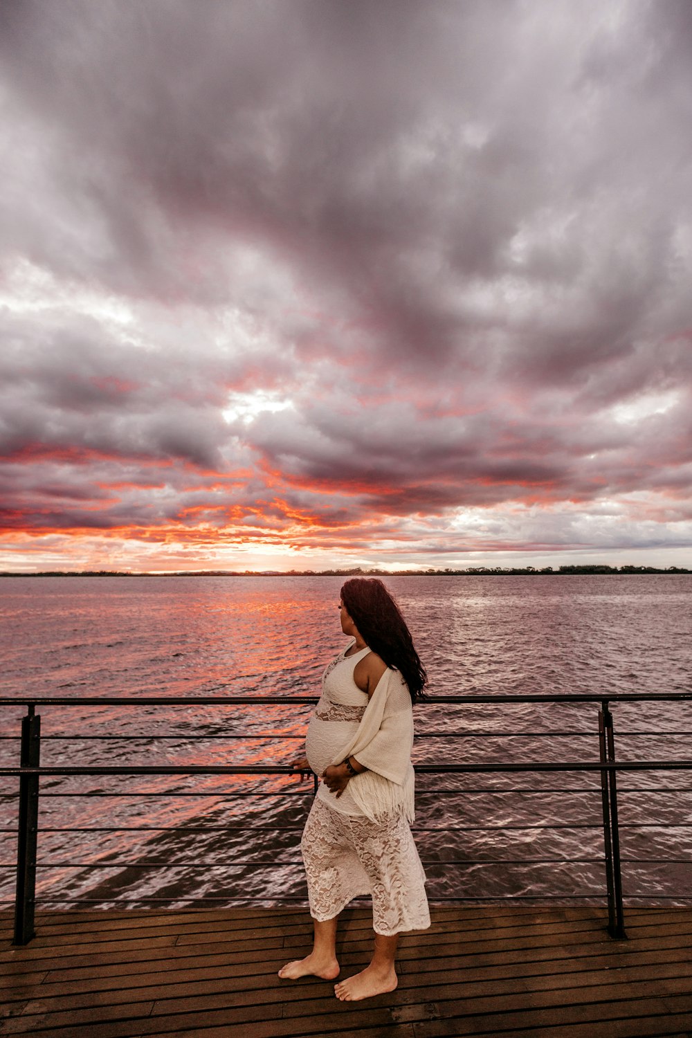 woman in white crop top and pants standing enar rail facing sea under heavy clouds