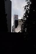 A tall building with a sign displaying 'PS' on the rooftop stands against a cloudy sky. The structure is framed by silhouettes of leaves and branches in the foreground. The building has an urban, industrial feel with antennas and other equipment on top.