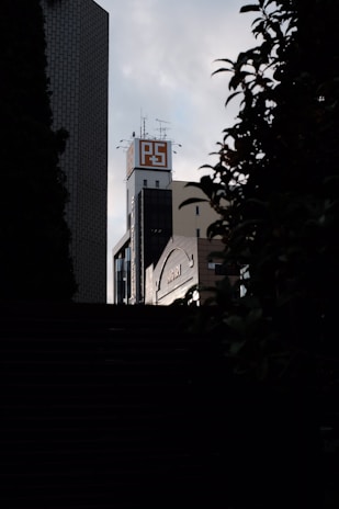 A tall building with a sign displaying 'PS' on the rooftop stands against a cloudy sky. The structure is framed by silhouettes of leaves and branches in the foreground. The building has an urban, industrial feel with antennas and other equipment on top.
