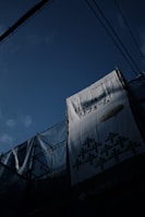 A large billboard with Japanese text hangs on a construction site, partially covered with scaffolding and netting. The sky is a deep blue, and electrical wires are visible overhead.