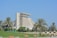 A large, modern hotel with a distinctive architectural design, surrounded by palm trees and a well-maintained green park. The sky is clear and blue, enhancing the serene and luxurious atmosphere. Benches and small structures are visible in the foreground, contributing to the inviting and relaxing environment.