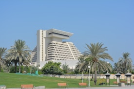 A large, modern hotel with a distinctive architectural design, surrounded by palm trees and a well-maintained green park. The sky is clear and blue, enhancing the serene and luxurious atmosphere. Benches and small structures are visible in the foreground, contributing to the inviting and relaxing environment.