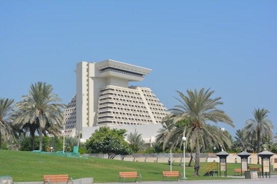 A large, modern hotel with a distinctive architectural design, surrounded by palm trees and a well-maintained green park. The sky is clear and blue, enhancing the serene and luxurious atmosphere. Benches and small structures are visible in the foreground, contributing to the inviting and relaxing environment.