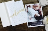 A wooden surface holds an open card with 'welcome' written in golden script alongside an announcement card. The announcement card features a photograph of an adult and a baby lying closely, with content expressions, and includes birth details. Nearby, a decorative box and white flowers add to the arrangement.