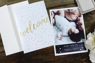 A wooden surface holds an open card with 'welcome' written in golden script alongside an announcement card. The announcement card features a photograph of an adult and a baby lying closely, with content expressions, and includes birth details. Nearby, a decorative box and white flowers add to the arrangement.