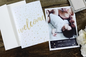 A wooden surface holds an open card with 'welcome' written in golden script alongside an announcement card. The announcement card features a photograph of an adult and a baby lying closely, with content expressions, and includes birth details. Nearby, a decorative box and white flowers add to the arrangement.