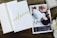 A wooden surface holds an open card with 'welcome' written in golden script alongside an announcement card. The announcement card features a photograph of an adult and a baby lying closely, with content expressions, and includes birth details. Nearby, a decorative box and white flowers add to the arrangement.