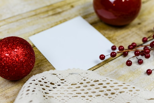 A wooden surface decorated with a small blank white card, a red glittery ornament, a piece of lace fabric, and a sprig of red berries. Two round red objects, one shiny and one matte, are positioned around the card.