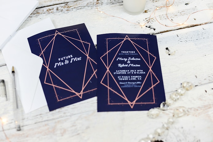 Wedding invitations are placed on a rustic white wooden table. The invitations feature a navy blue background with elegant geometric designs in rose gold. One card displays the words 'Future Mr & Mrs,' while the other provides details about the wedding, including names and the date. Nearby are a pen, an envelope, and some string lights, creating a warm and inviting atmosphere.